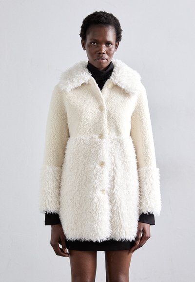 Manteau blanc et duveteux en matière douce ; il possède un grand col texturé, quatre boutons et des manches longues avec une texture pelucheuse.