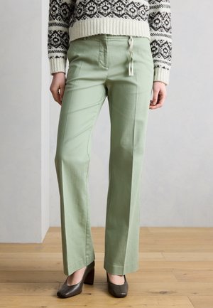 Osoba v svetlozelených nohaviciach s rovnými nohavicami, čiernych lodičkách s podpätkom a bielom a čiernom vzorovanom svetri stojaca na drevenej podlahe.