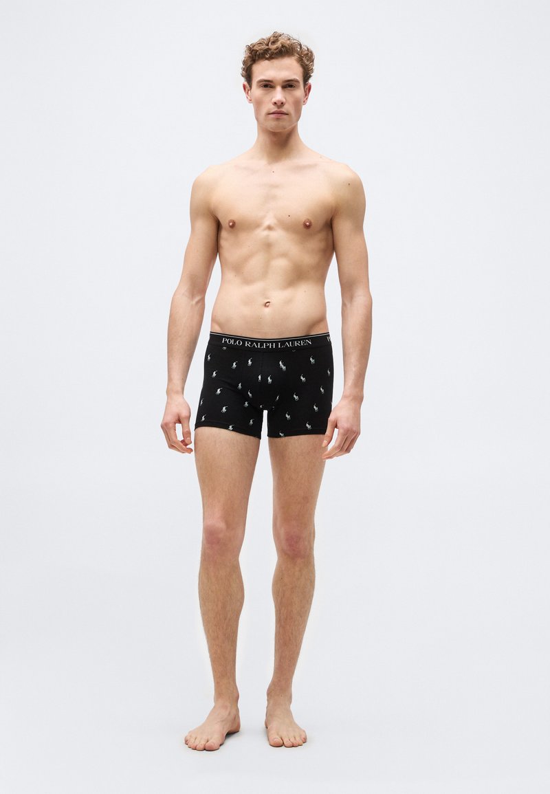 Νέος άντρας χωρίς πουκάμισο, όρθιος ξυπόλητος, φ οράει μαύρα εσώρουχα Polo Ralph Lauren με λευκά λογότυπα σε απλό ανοιχτό φόντο.