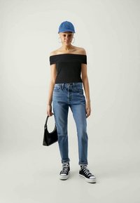 Top preto de ombros à mostra, jeans azuis de cintura alta, ténis pretos e um boné azul, segurando uma pequena bolsa preta.