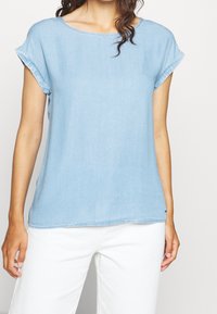 Ljust blå denimtop med korta uppvikta ärmar, rundad halsringning och en avslappnad passform. Mjuk textur med synliga sömdetaljer längs fållen.