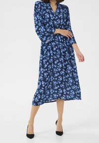 Navy sininen kukallinen mekko, jossa on vaaleansinisiä kukkia, V-pääntie, pitkät hihat ja kapea vyötärö, saavuttaen puoliväliä sääristä, yhdistettynä mustiin korkokenkiin.