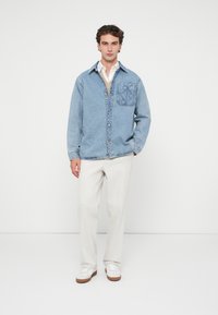 Camisa de ganga de botão em azul claro com um bolso no peito e costura contrastante, combinada com calças creme e ténis brancos.