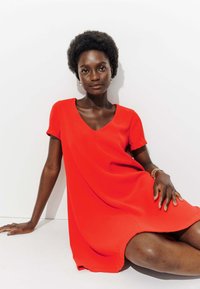 Robe rouge à manches courtes avec un décolleté en V, fabriquée en tissu lisse, coupe drapée, se terminant au-dessus du genou. Modèle assis sur un fond blanc.