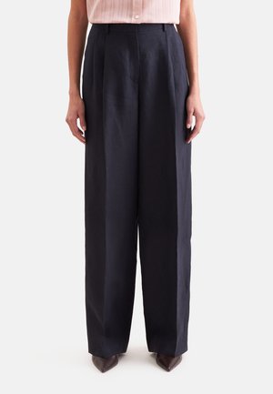 Femme portant un pantalon noir taille haute à jambes larges, associé à des chaussures pointues marron et un chemisier rose clair rentré.