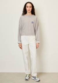 Siva majica z dolgimi rokavi z majhnim modrim logotipom, v kombinaciji z belimi hlačami z visokim pasom in sivimi športnimi čevlji, ki stoji ob nevtralni ozadju.