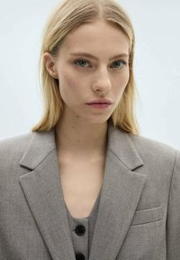 Blazer gris sur mesure avec revers, doté d'une poche unique. En dessous, un haut gris à col en V avec fermeture à boutons. Texture lisse.