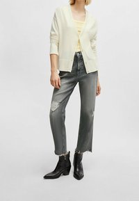 Ljus krämfärgad cardigan med knappar, lager på en blekgul topp. Grå slitna jeans med fransade fållar och svarta ankelskor.