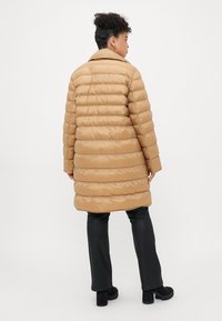 Kvinne som har på seg en lang beige pufferjakke, svarte bukser og svarte høyhælte støvler, stående med ryggen mot en enkel hvit bakgrunn.