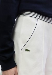 Pantalon blanc texturé avec ceinture rayée marine, présentant un logo crocodile vert brodé sur le côté. Manches longues bleu marine visibles.