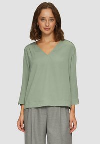 Blouse vert clair à col en V avec des manches trois-quarts, tissu lisse et détails texturés subtils le long des épaules. Associée à un pantalon gris.