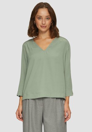 Blouse vert clair à col en V avec des manches trois-quarts, tissu lisse et détails texturés subtils le long des épaules. Associée à un pantalon gris.