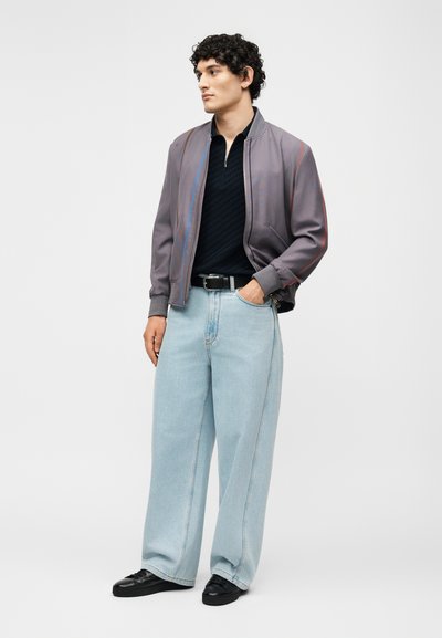 Άνδρας με σγουρά μαλλιά φοράει γκρι μπουφάν bomber, μαύρο πουλόβερ, ανοιχτό μπλε παντελόνι με φαρδιές μπατζάκια, μαύρα παπούτσια και μαύρη ζώνη, στέκεται με το ένα χέρι στην τσέπη.