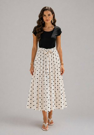 Femme portant un haut noir à manches courtes et une jupe blanche longue jusqu'aux chevilles avec des pois noirs et une ceinture nouée à la taille, debout pieds nus.