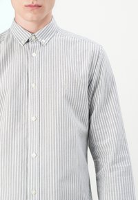 Stribet button-up skjorte i lysegrå og hvid. Har spredt krave, lange ærmer og en subtil broderet logo på brystet.