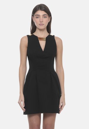 Donna che indossa un vestito nero senza maniche con scollo a V e dettagli metallici dorati sulle spalle e sullo scollo, in piedi davanti a uno sfondo semplice.