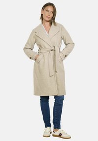 Bežinis vilnos mišinio tranšėjaus paltas su diržu, įpjautomis apykaklėmis ir prisiūtomis rankovėmis, derinamas su tamsiai mėlynomis džinsomis ir šviesiais sportbačiais.