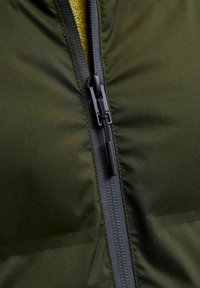 Zbliżenie na zamek błyskawiczny zielonej kurtki z czarną zawieszką oraz widoczną żółtą dzianiną pod spodem, ukazujące fakturę materiału i ścieg.