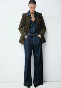Pantaloni a gamba larga in denim blu scuro abbinati a un blazer in velluto verde scuro e una camicia in denim coordinata, con design a bottoni e cintura.