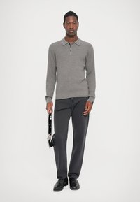 Grijze geribde gebreide polo trui met een kraag, gecombineerd met donkergrijze broeken. Model houdt een zwarte tas vast en draagt zwarte schoenen. Effen achtergrond.