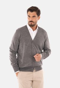 Cardigan grigio in lana con scollatura a V, chiusura a bottoni e polsini a costine. Indossato sopra una camicia bianca, sorretto da un modello su uno sfondo chiaro.