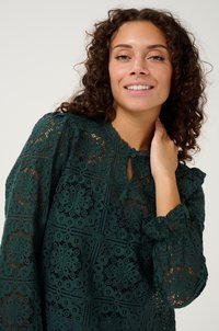 Dark green lace top with floral patterns, keyhole neckline, frilled cuffs, and textured design. Lightweight and semi-transparent fabric.