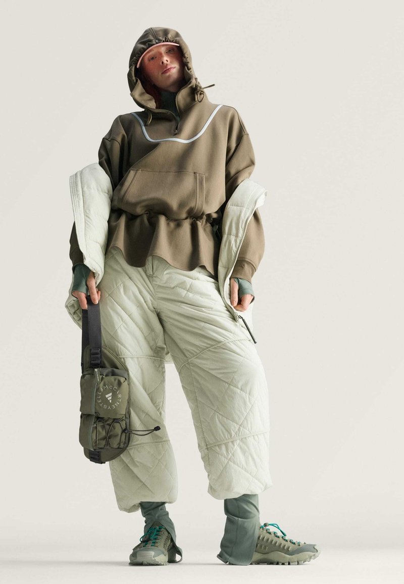 Olivgrüner Kapuzenpullover mit hohem Kragen und verstellbarer Kapuze, kombiniert mit hellgrünen gesteppte Hosen. Das Outfit hat eine strukturierte Oberfläche und passende Schuhe.