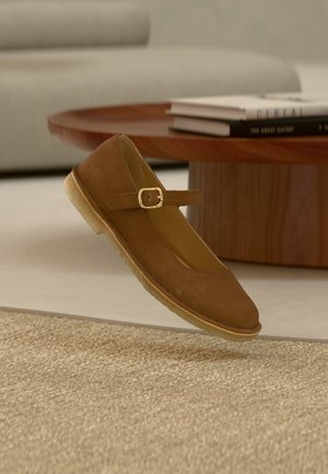 Chaussure Mary Jane marron avec boucle dorée flottant au-dessus d'un tapis beige texturé, table basse ronde en bois et livres en arrière-plan.