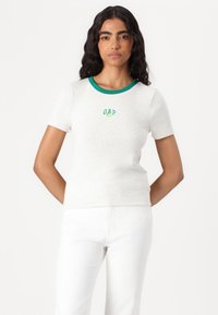 Camiseta gris de manga corta con cuello acanalado verde y la palabra "GAP" bordada en verde. Textura suave, diseño ajustado, combinada con pantalones blancos.