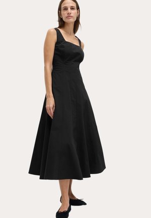 Femme portant une robe midi noire sans manches, cintrée avec une jupe évasée et un décolleté carré, associée à des ballerines noires.