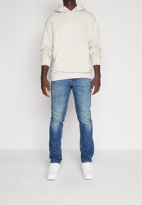 Felpa con cappuccio beige chiaro con tasca frontale, abbinata a jeans di un blu medio. Il modello indossa scarpe da ginnastica bianche. Design semplice e casual, con una vestibilità comoda.