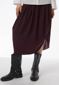 Jupe midi bordeaux avec taille froncée, dotée d'une fente sur le côté ; associée à des bottes noires en cuir jusqu'aux genoux avec des accents de boucles.