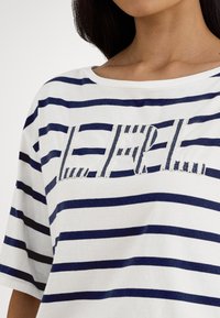 Camisola branca de algodão com riscas verticais azuis marinho, corte descontraído e um logótipo texturizado "L.E.F.L." no centro.