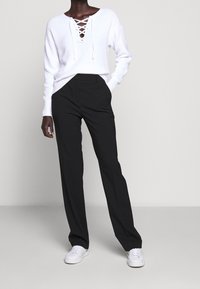 Maglione bianco a maglia con scollatura profonda a V e dettagli con lacci, abbinato a pantaloni neri eleganti e sneakers bianche. Design semplice e funzionale.