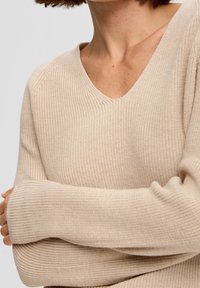 Pull beige en tricot avec un col en V, une texture côtelée et des manches longues, présentant une apparence douce et confortable.