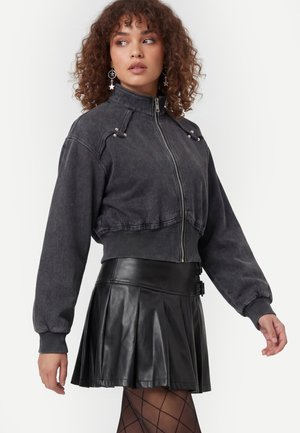 Donna con capelli ricci che indossa orecchini a forma di stella, una giacca corta scura con zip, una gonna nera di pelle plissettata e collant a fantasia.