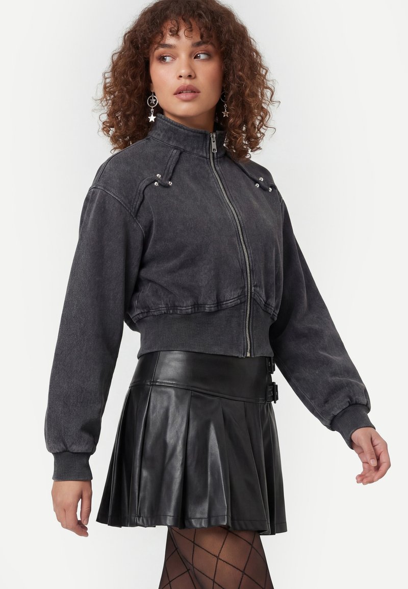 Femme aux cheveux bouclés portant des boucles d’oreilles en forme d’étoile, une veste courte foncée zippée, une jupe plissée en cuir noire et des collants à motifs.