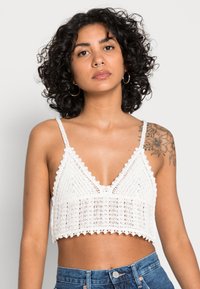 Bralette de crochê branco com decote em V, bordas em forma de onda e design cropped. A textura é de malha aberta com um padrão geométrico.