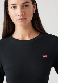 Crna majica s dugim rukavima od rebraste tkanine, s malim crvenim Levi's logom na lijevoj strani prsnog područja. Jednostavan, okrugli izrez.