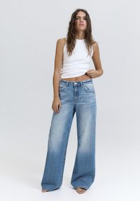 Kobieta o falowanych włosach, ubrana w białą, bezrękawnikową bluzkę i szerokie, wyblakłe niebieskie dżinsy z małymi białymi plamkami, stojąca boso.