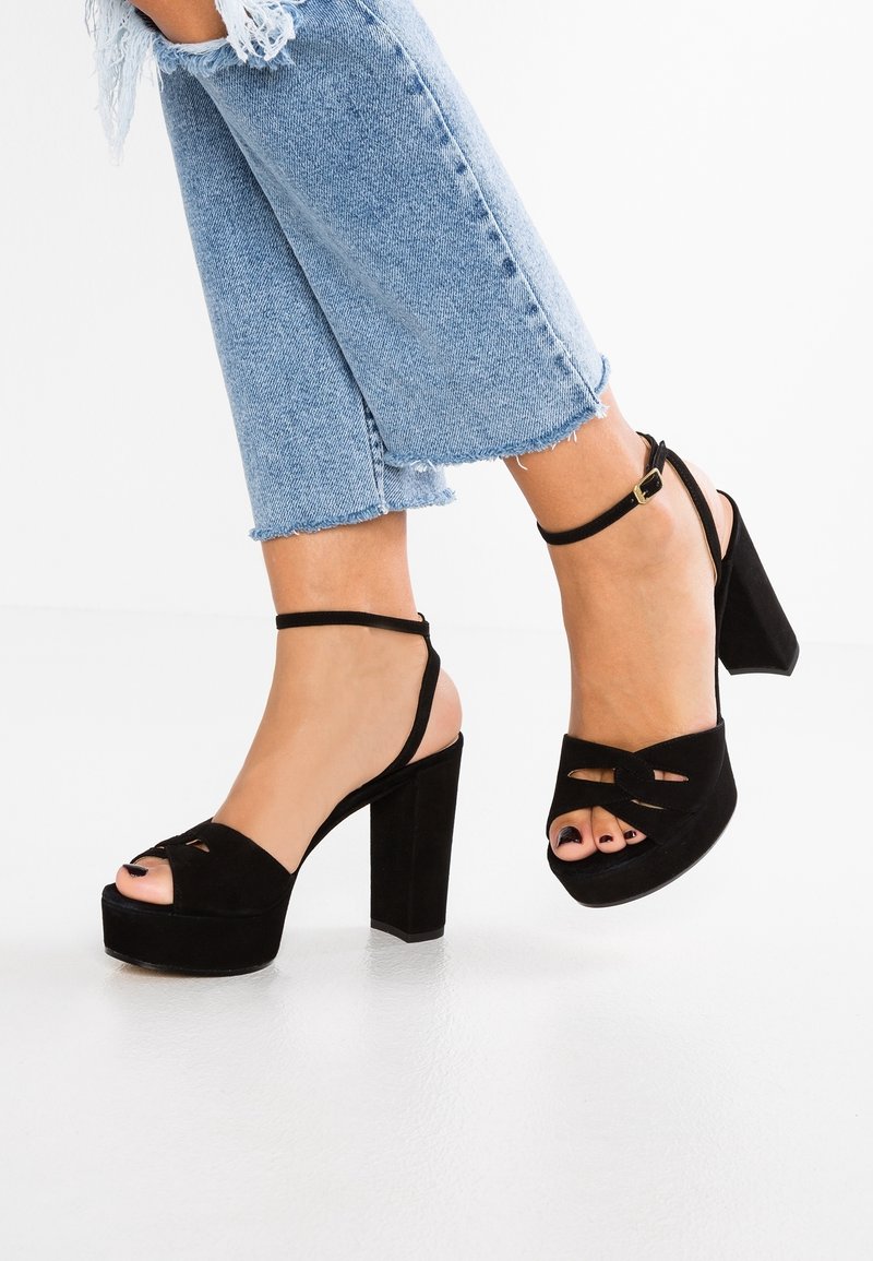 Pieds portant des sandales à talons hauts à plateforme noires avec des brides à la cheville, associées à un jean court bleu à ourlet effiloché sur un sol blanc.