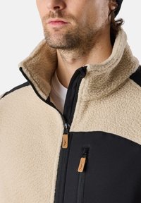 Fleece jacket featuring a beige body, black panels, high collar, and two zippered pockets with leather accents. Soft texture throughout.