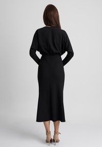 Czarna midi sukienka z długimi rękawami, elastyczną talią i rozcięciem z tyłu. Gładki materiał o delikatnym połysku. Noszona z beżowymi szpilkami.