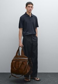 Svart polo-tröja med korta ärmar, randiga byxor och en brun mockaväska med silverdetaljer och justerbara remmar. Flip-flops på fötterna.