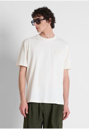 Uomo che indossa una maglietta bianca oversize con "AM" impresso sul petto e pantaloni verde scuro, guarda di lato indossando grandi occhiali da sole neri.