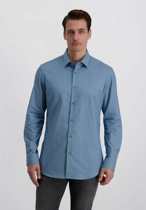 Mann trägt ein langärmliges hellblaues Hemd mit Knöpfen und dunkle Hose, steht vor einem schlichten hellen Hintergrund.