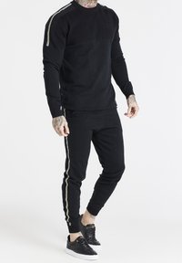 Svart sweatshirt och matchande joggers med gyllene sidostråkar, tillverkade av mjukt tyg. Har rundad hals och ribbade handledsmuddar. Svarta sneakers.
