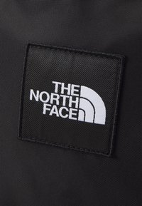 Patch en tissu noir avec texte brodé blanc "THE NORTH FACE" et logo en demi-dôme cousu sur une surface texturée noire.