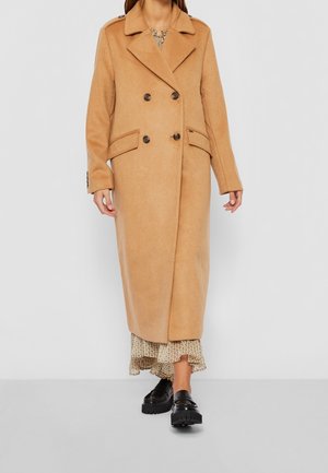 Kobieta ubrana w długi, dwurzędowy płaszcz w kolorze wielbłądzim, na kwiecistej sukience, z czarnymi masywnymi mokasynami, stojąca na jednolitym tle.