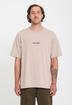 Homme aux cheveux mi-longs portant un t-shirt beige Volcom et un pantalon olive, debout devant un fond blanc uni.